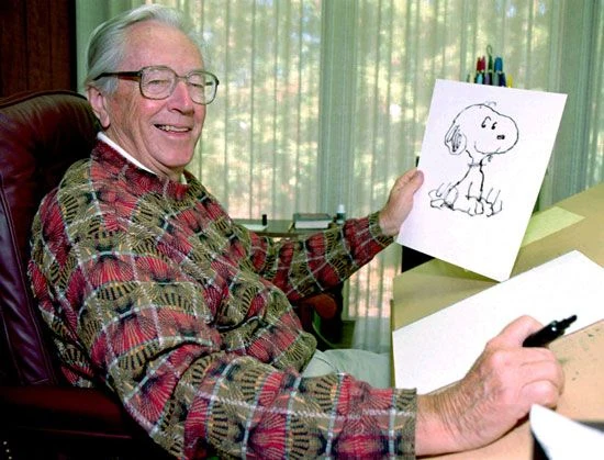 Charles M. Schulz smiling at his drawing board and holding a drawing of Snoopy